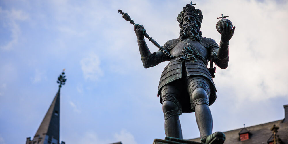 Bronzestatue Karls des Großen auf dem Karlsbrunnen in Aachen, mit Krone, Zepter und Reichsapfel vor blauem Himmel.