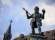 Bronzestatue Karls des Großen auf dem Karlsbrunnen in Aachen, mit Krone, Zepter und Reichsapfel vor blauem Himmel.