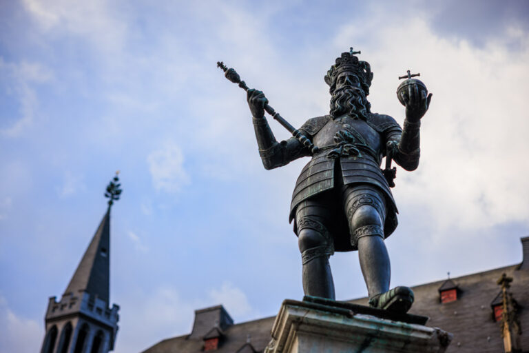 Bronzestatue Karls des Großen auf dem Karlsbrunnen in Aachen, mit Krone, Zepter und Reichsapfel vor blauem Himmel.