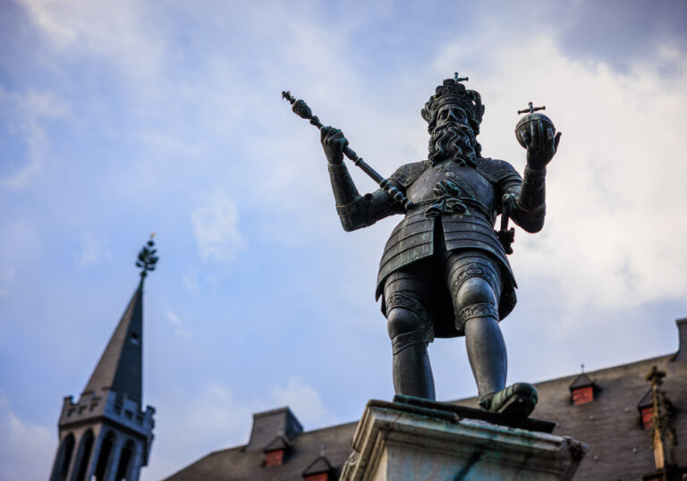 Bronzestatue Karls des Großen auf dem Karlsbrunnen in Aachen, mit Krone, Zepter und Reichsapfel vor blauem Himmel.