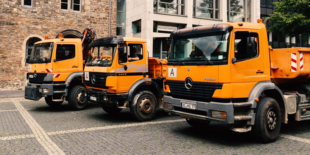 Orangefarbene Mercedes-Kipplaster parken auf dem Pflaster vor einem historischen Sandsteingebäude nahe dem Aachener Dom.