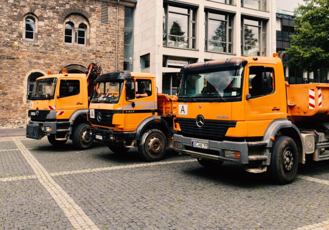 Orangefarbene Mercedes-Kipplaster parken auf dem Pflaster vor einem historischen Sandsteingebäude nahe dem Aachener Dom.