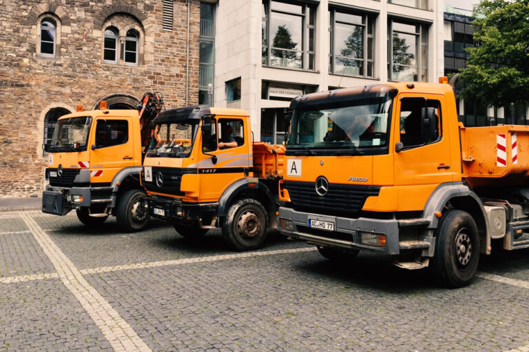 Orangefarbene Mercedes-Kipplaster parken auf dem Pflaster vor einem historischen Sandsteingebäude nahe dem Aachener Dom.