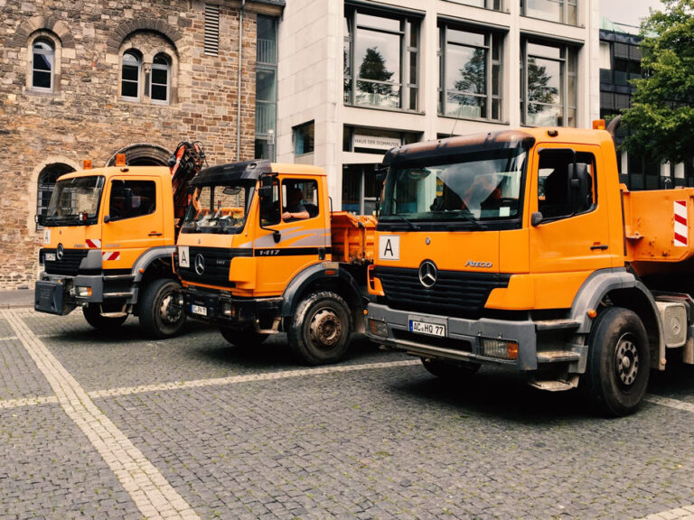 Orangefarbene Mercedes-Kipplaster parken auf dem Pflaster vor einem historischen Sandsteingebäude nahe dem Aachener Dom.