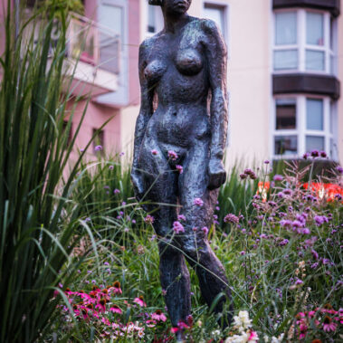 Bronzeskulptur einer stehenden weiblichen Figur (Mädchenplastik von Schepp) inmitten eines Blumenbeets in Burtscheid, Wohnhäuser im Hintergrund