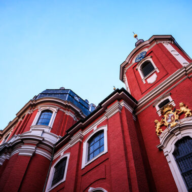 Untersicht der barocken Fassade der Kirche St. Michael in Burtscheid, rotes Backsteinmauerwerk mit goldenem Wappen und Löwenfiguren, Uhrturm