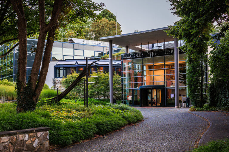 Gläserner Eingangsbereich der Carolus Thermen Aachen mit Schriftzug, umgeben von Grünanlagen