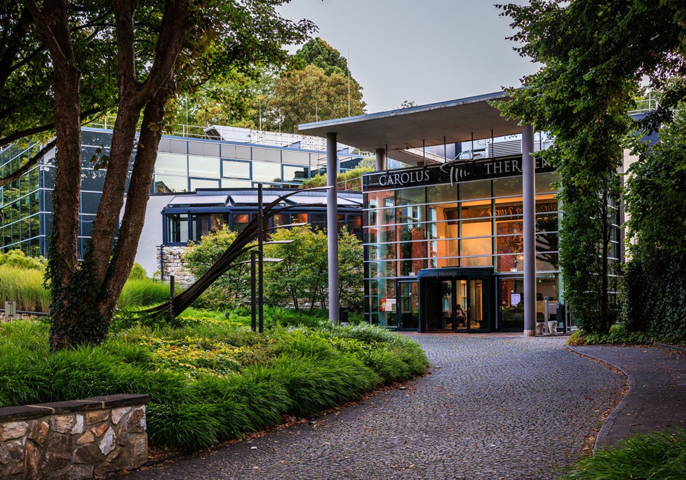 Gläserner Eingangsbereich der Carolus Thermen Aachen mit Schriftzug, umgeben von Grünanlagen