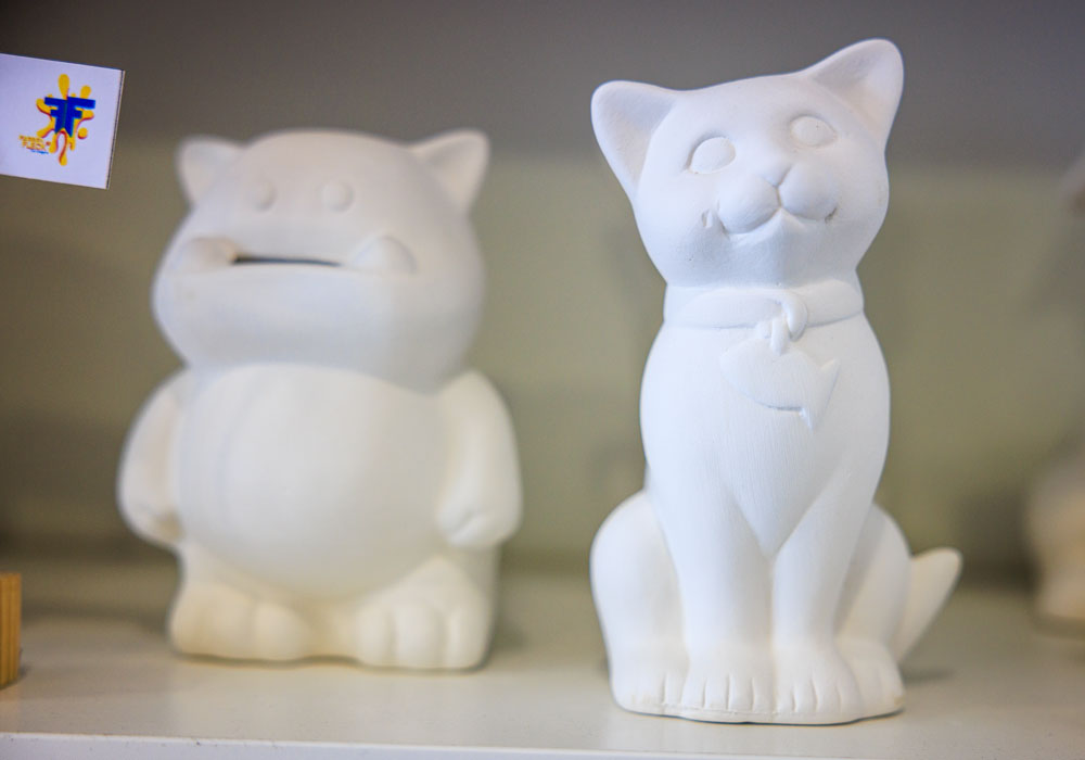 Zwei unbemalte weiße Keramikfiguren im Regal bei Farbenfleck Aachen, eine sitzende Katze und eine runde Spardosen-Figur, bereit zum Bemalen