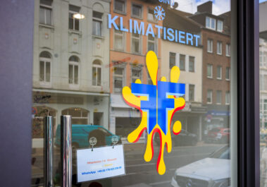 Schaufenster des Keramik-Malstudios Farbenfleck Aachen mit gelb-blauem Logo, Hinweis auf Klimatisierung und Stellenanzeige, Spiegelung der gegenüberliegenden Häuserzeile