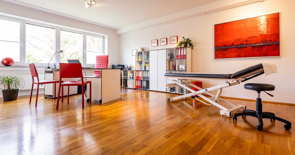 Behandlungsraum der Hausarztpraxis Sailer mit Liege, Schreibtisch und rotem Wandbild in hellem, freundlichem Ambiente