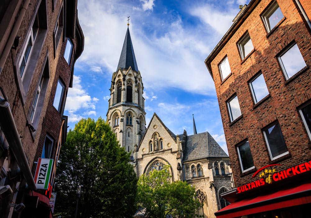 Turm der Heilig-Kreuz-Kirche über den Hausdächern der Pontstraße, umrahmt von Imbiss- und Pizzaschildern