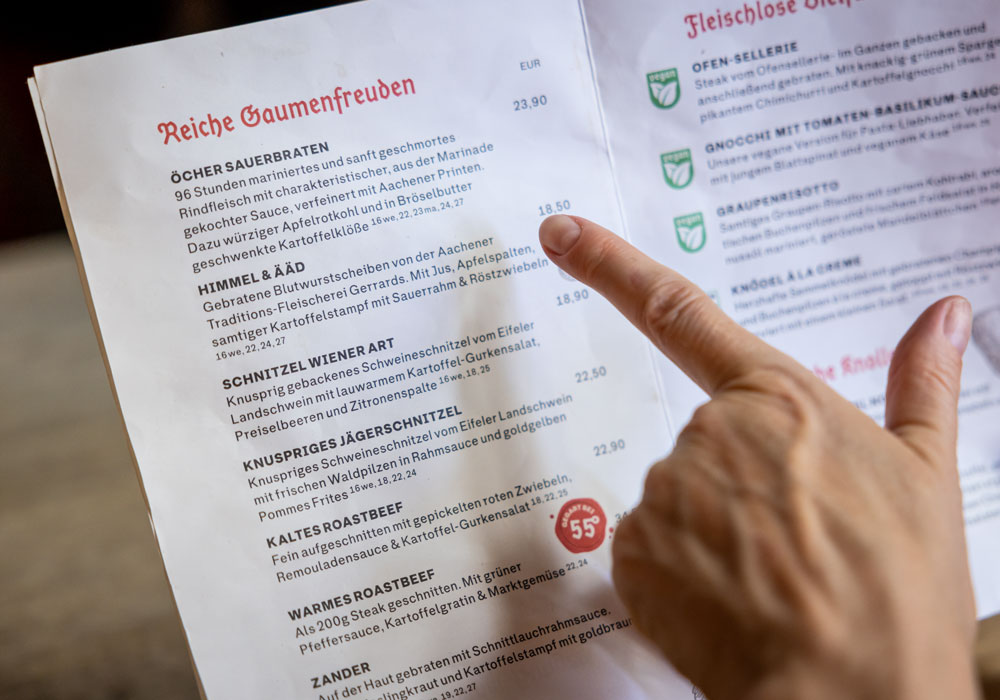 Speisekarte des Postwagen Zum Ratskeller, ein Finger zeigt auf den Eintrag „Himmel & Ääd" in der Rubrik Reiche Gaumenfreuden
