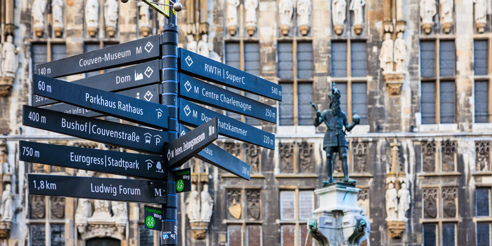 Wegweiser mit Richtungsschildern zu Aachener Sehenswürdigkeiten vor der Rathausfassade, rechts die Karlsbrunnen-Statue