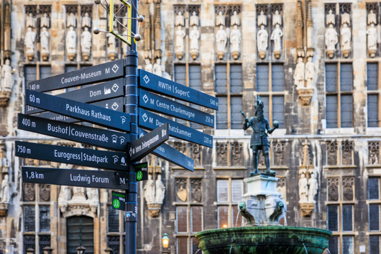 Wegweiser mit Richtungsschildern zu Aachener Sehenswürdigkeiten vor der Rathausfassade, rechts die Karlsbrunnen-Statue
