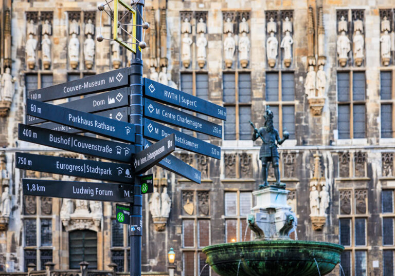 Wegweiser mit Richtungsschildern zu Aachener Sehenswürdigkeiten vor der Rathausfassade, rechts die Karlsbrunnen-Statue