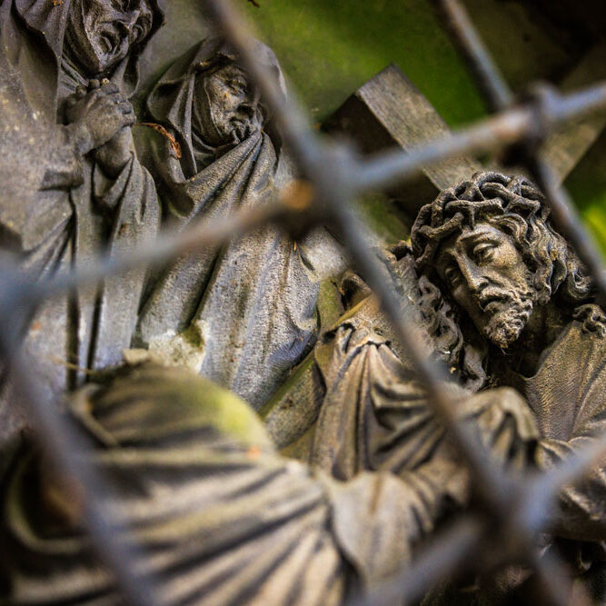 Steinrelief eines Kreuzwegs auf dem Lousberg, durch ein Gitter fotografiert, zeigt Jesus mit Kreuz und Dornenkrone