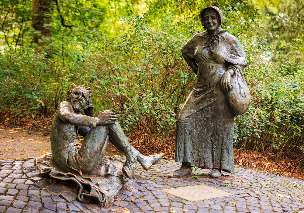 Bronzestatuen von Bauersfrau und Teufel am Denkmalplatz auf dem Lousberg, umgeben von herbstlichem Grün