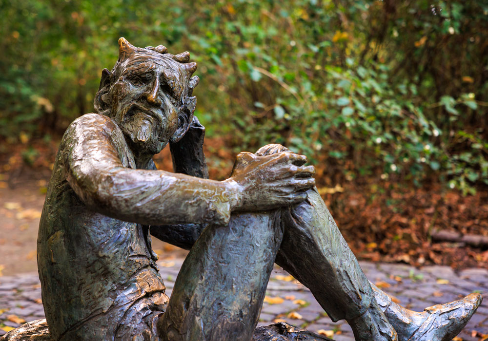 Nahaufnahme der Bronzestatue des erschöpften Teufels am Lousberg-Denkmal, sitzend mit Hörnern und nachdenklichem Gesichtsausdruck