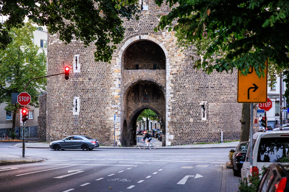 Marschiertor Aachen – Straßenansicht Das Aachener Marschiertor an einer belebten Kreuzung mit Ampel, Stoppschildern und einem Radfahrer, der durch den Torbogen fährt