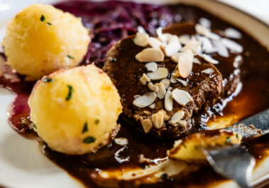 Nahaufnahme des Öcher Sauerbratens: geschmorte Rinderscheibe mit Mandelblättchen in dunkler Printensoße, Kartoffelkloß und Apfelrotkohl, Gabel am Tellerrand