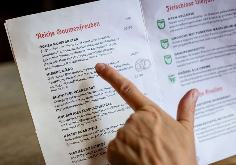Speisekarte des Postwagen Zum Ratskeller, zwei Finger zeigen auf die Einträge „Himmel & Ääd" und „Schnitzel Wiener Art" in der Rubrik Reiche Gaumenfreuden