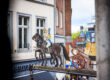 Historisches Zunftschild des Postwagens in Aachen: bemalte Postkutsche mit Pferden und Postillion vor Altstadthäusern, im Hintergrund das Geschäft Ghorran Delikatessen