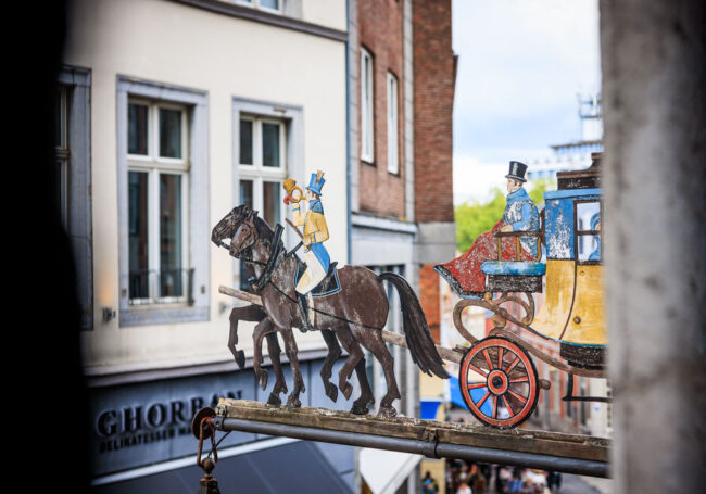 Historisches Zunftschild des Postwagens in Aachen: bemalte Postkutsche mit Pferden und Postillion vor Altstadthäusern, im Hintergrund das Geschäft Ghorran Delikatessen