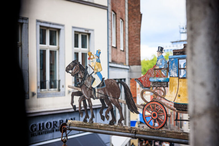 Historisches Zunftschild des Postwagens in Aachen: bemalte Postkutsche mit Pferden und Postillion vor Altstadthäusern, im Hintergrund das Geschäft Ghorran Delikatessen