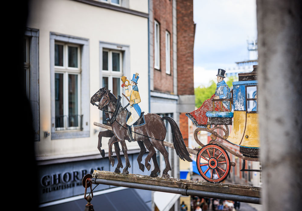 Historisches Zunftschild des Postwagens in Aachen: bemalte Postkutsche mit Pferden und Postillion vor Altstadthäusern, im Hintergrund das Geschäft Ghorran Delikatessen