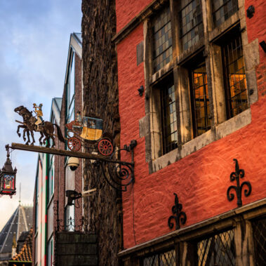 Historisches Postwagen-Schild an der roten Fassade am Aachener Markt, bemalte Postkutsche mit Pferden, schmiedeeiserne Laterne, Abendhimmel im Hintergrund
