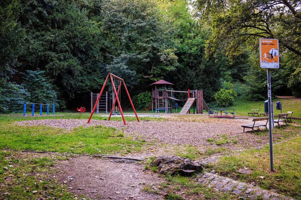 Übersicht des Spielplatzes Buchenallee mit Schaukel, Klettergerüst und Spielturm, daneben ein Hinweisschild „Spielplatz Buchenallee"