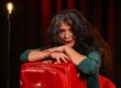 Sanaz Zaresani lehnt an einem roten Koffer, im Hintergrund eine Glühbirnenlampe und roter Vorhang