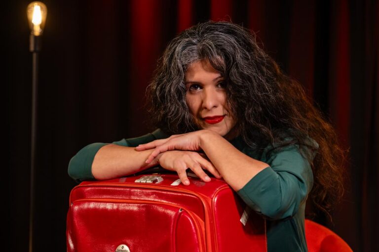 Sanaz Zaresani lehnt an einem roten Koffer, im Hintergrund eine Glühbirnenlampe und roter Vorhang