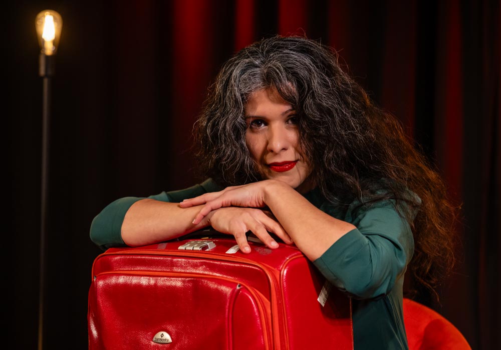 Sanaz Zaresani lehnt an einem roten Koffer, im Hintergrund eine Glühbirnenlampe und roter Vorhang