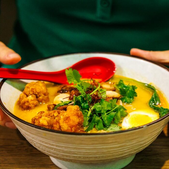 Creamy White Tan Tan Men bei Takumi Aachen mit frittiertem Hähnchen, halbem Ei und frischem Koriander, mit beiden Händen präsentiert