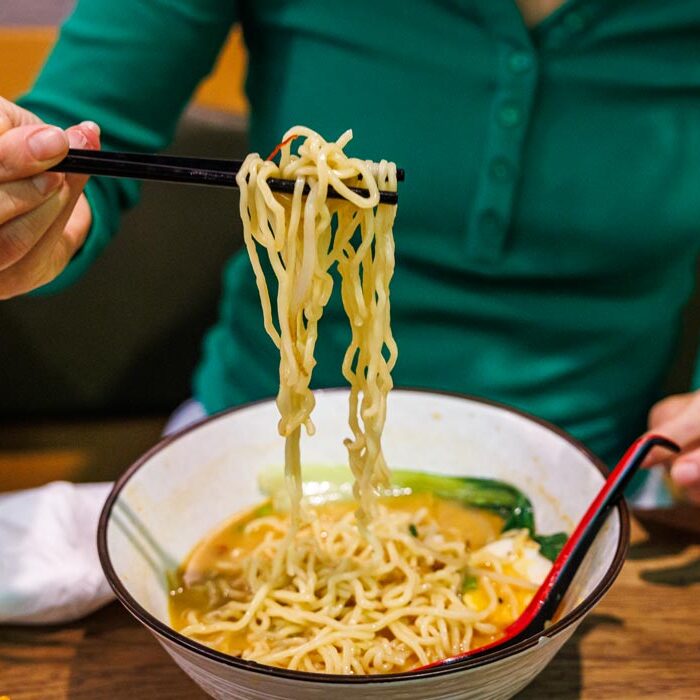 Ramen-Nudeln werden mit Stäbchen aus der Schüssel gehoben, im Hintergrund die Creamy White Tan Tan Men bei Takumi Aachen