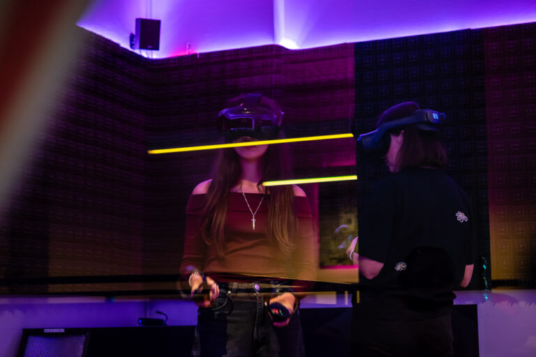 Zwei Spielerinnen mit VR-Headsets in violett beleuchteter Arena, durch eine Glasscheibe mit Spiegelungen fotografiert