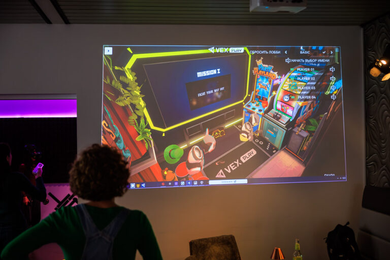 Spiellobby eines VR-Spiels auf der Leinwand im Blauwal VR-Café Aachen, davor eine Zuschauerin